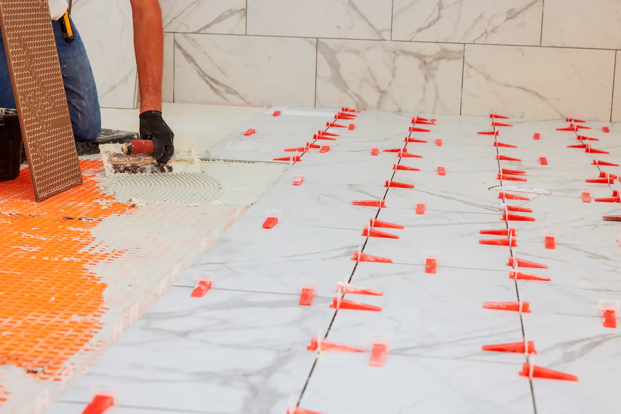 Person installing large marble tiles on a floor using orange spacers and adhesive beside a tiled wall.